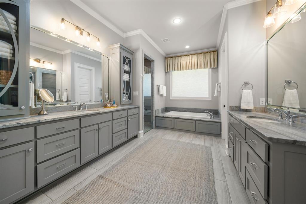 Dual gray cabinetry vanities with stone countertops, undermount sinks, full-width mirrors, deep soaking tub, and tile flooring