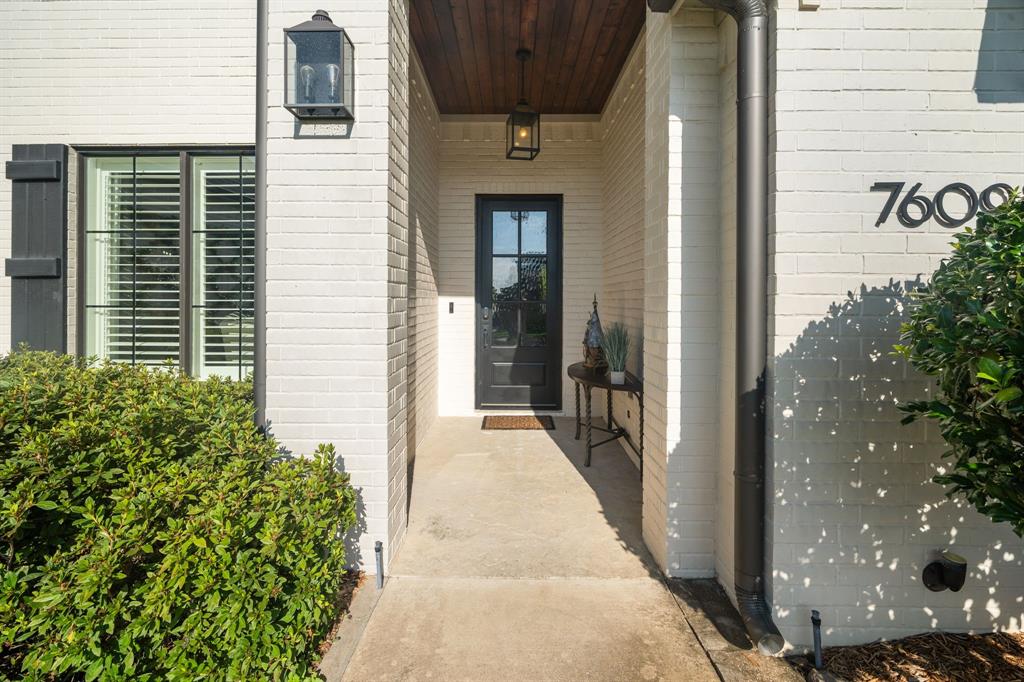 Covered entryway featuring a painted brick facade, wood-plank ceiling, black metal-framed front door with glass inlay, and structured exterior lantern lighting