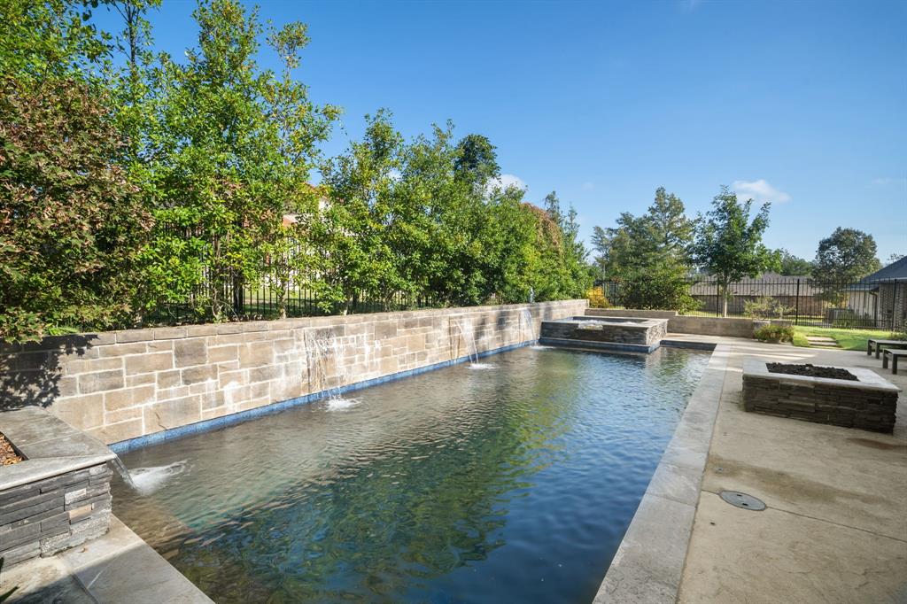 Outdoor area featuring a rectangular pool with integrated water features, raised stone retaining wall, stone-surround spa, and a stone fire pit set on a concrete patio Outdoor area featuring a rectangular pool with integrated water features, raised stone retaining wall, stone-surround spa, and a stone fire pit set on a concrete patio