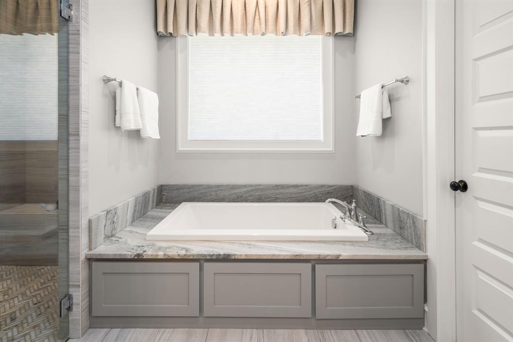 Soaking tub with stone surround, custom gray cabinetry, chrome deck-mounted faucet, and window with neutral window treatment