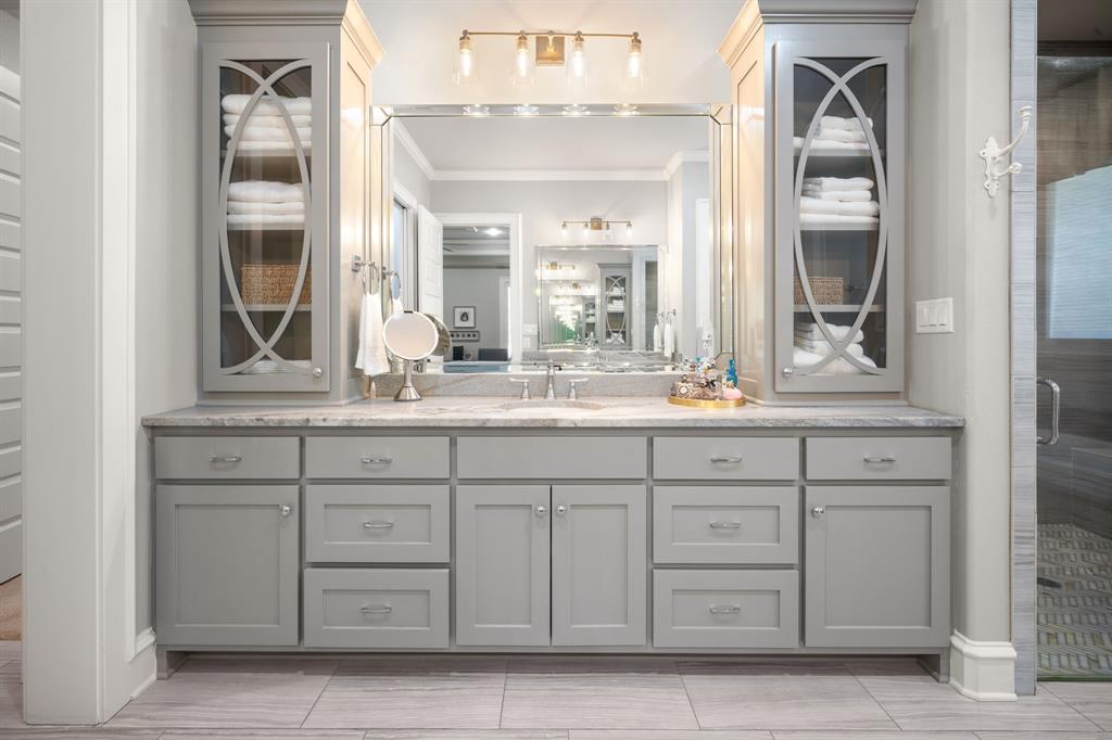 Dual-sink vanity featuring gray shaker-style cabinetry, integrated glass-front shelving, a natural stone countertop, and an oversized polished-edge mirror with multi-bulb vanity lighting
