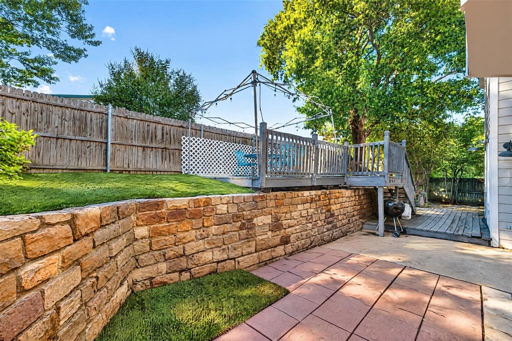 Tiered backyard featuring a stone retaining wall, paved patio area, raised wooden deck, and perimeter wood fencing