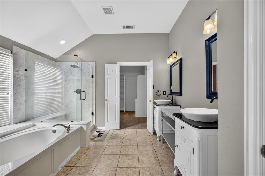 Dual vanities featuring white cabinetry, dark countertops, and vessel sinks, complemented by a glass-enclosed shower with stone-finish surround and a deep soaking tub