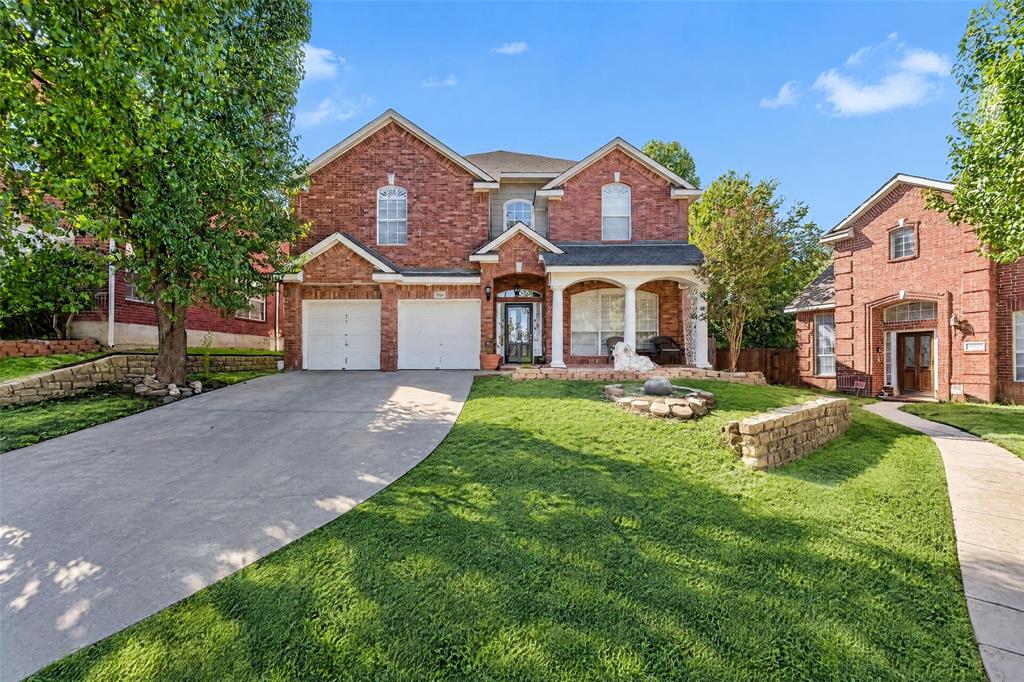 Two-story brick exterior featuring a multi-gabled roofline, covered front porch with arched columns, attached two-car garage, and stone hardscaping