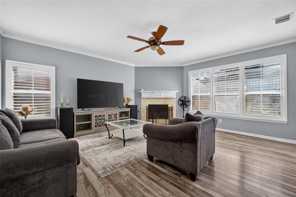 Living room featuring wood-finish flooring, a fireplace with a mantel, crown molding, a ceiling fan, and large windows with integrated blinds
