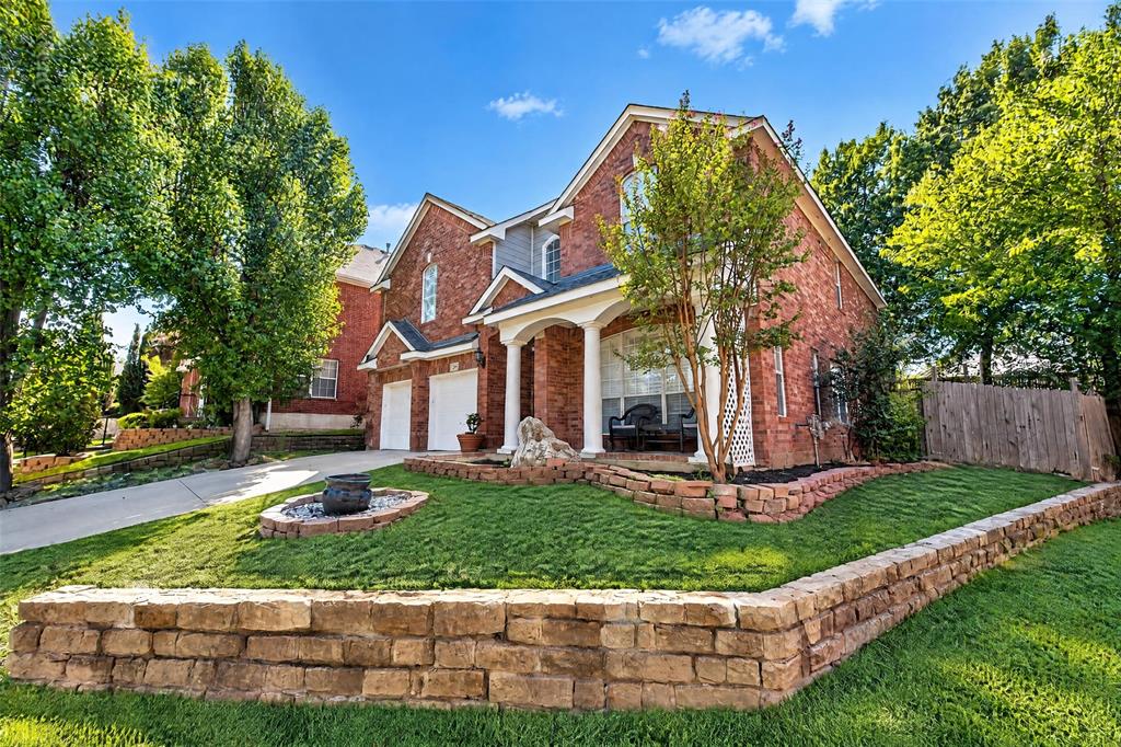 Traditional brick exterior featuring a covered front porch with white columns, two-car garage, and tiered natural stone retaining walls