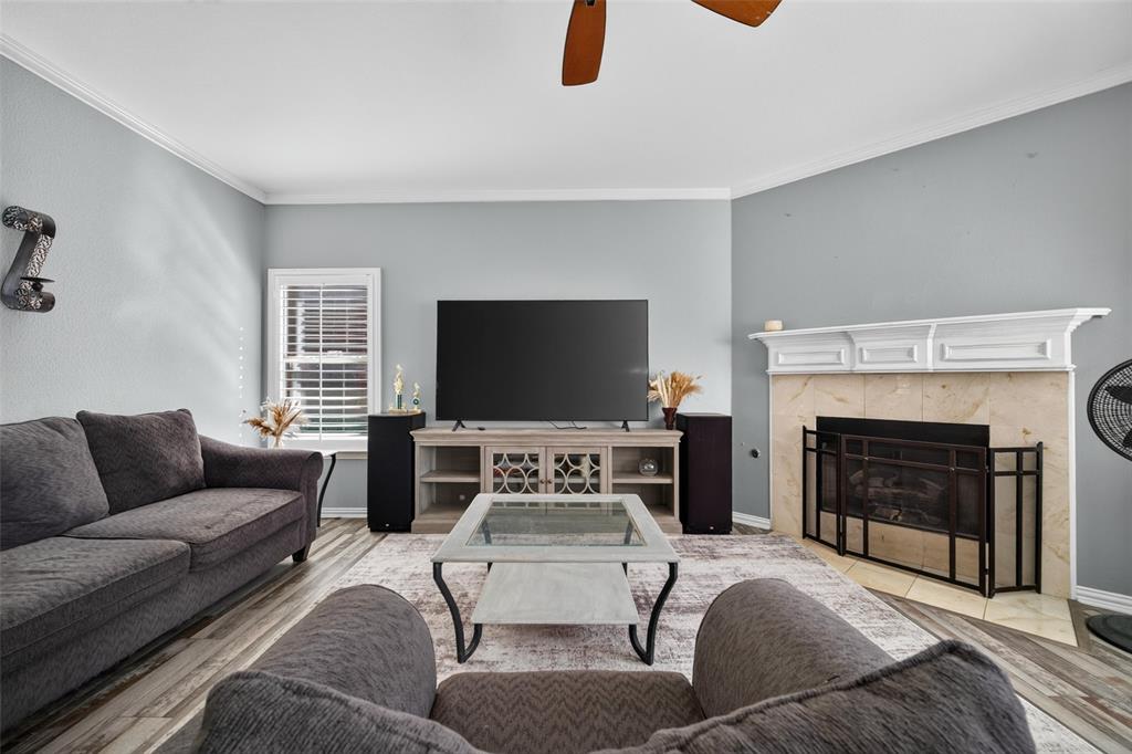Living area featuring wood-finish flooring, a fireplace with a stone-surround and white mantel, crown molding, plantation shutters, and a ceiling fan