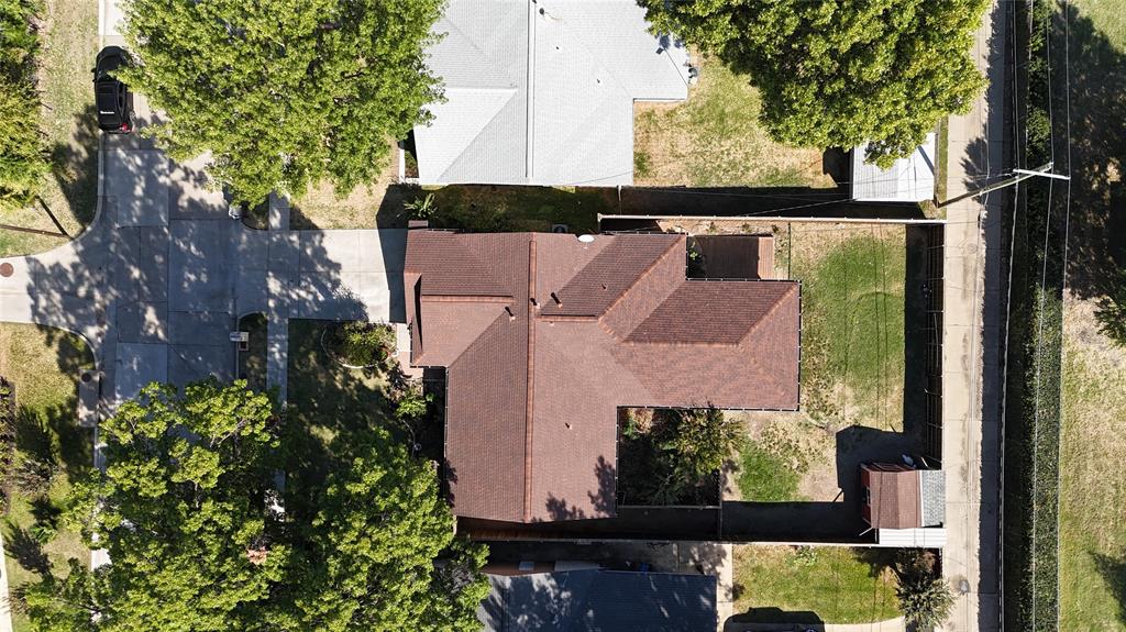 Aerial perspective featuring a multi-pitch shingle roof, concrete driveway, fenced perimeter, and mature canopy trees