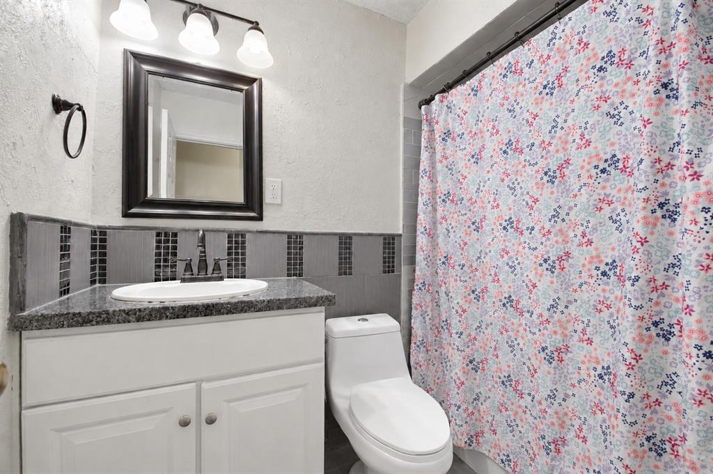 White vanity cabinet with a stone-finish countertop, oval undermount sink, dark-toned mosaic tile backsplash, and a three-light vanity fixture