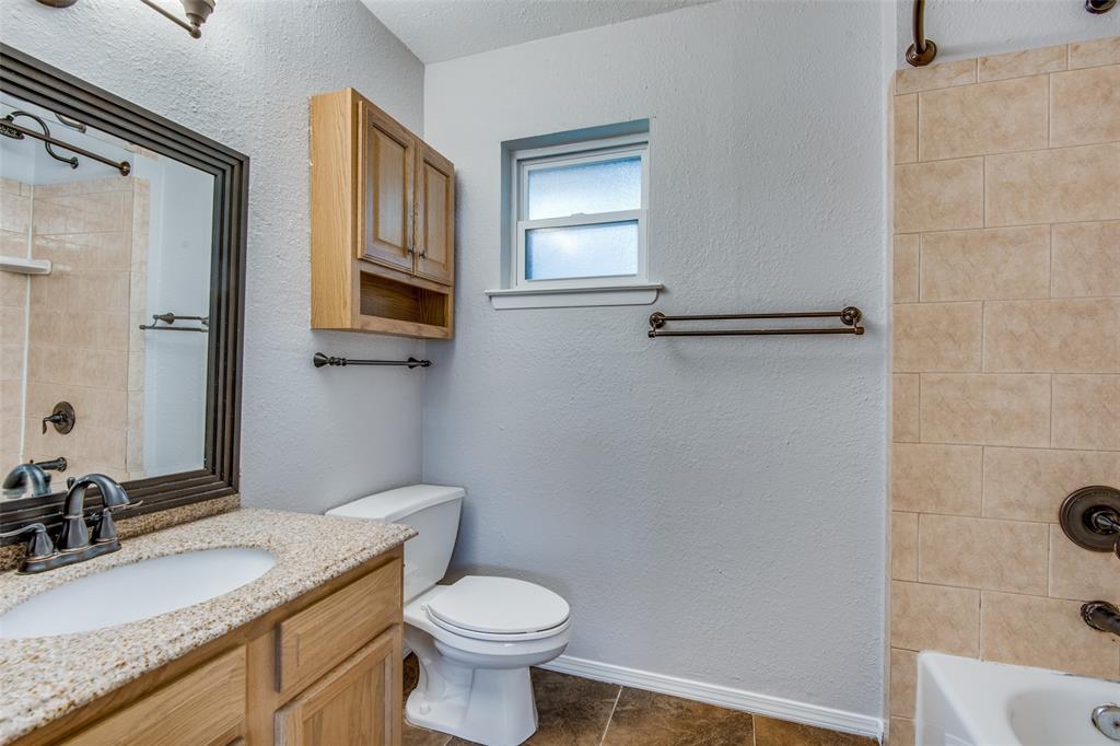 Bathroom featuring a vanity with a stone-finish countertop and an integrated sink, framed wall mirror, wood-finish wall-mounted cabinetry, a tiled tub surround, and dark-toned metal hardware Bathroom featuring a vanity with a stone-finish countertop and an integrated sink, framed wall mirror, wood-finish wall-mounted cabinetry, a tiled tub surround, and dark-toned metal hardware