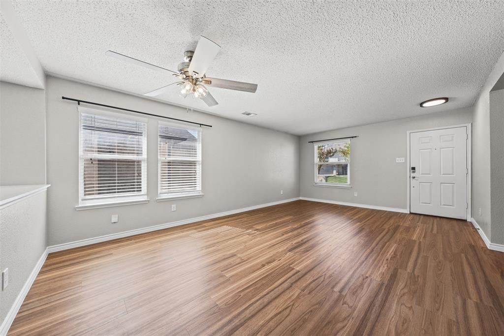 Spacious room featuring wood-finish flooring, ceiling fan with integrated lighting, dual-pane windows with horizontal blinds, and a paneled entry door Spacious room featuring wood-finish flooring, ceiling fan with integrated lighting, dual-pane windows with horizontal blinds, and a paneled entry door