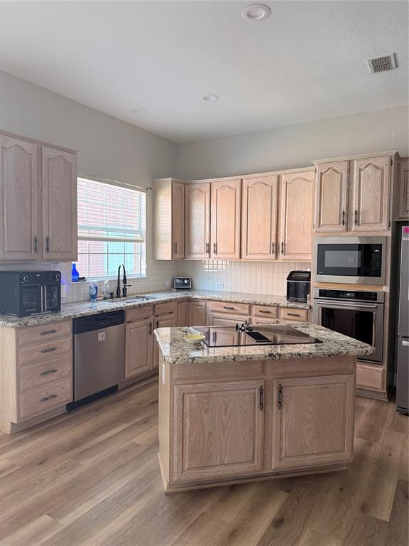 Kitchen featuring light-tone wood cabinetry, stone countertops, a central island with a built-in cooktop, stainless steel appliances, and wood-finish flooring