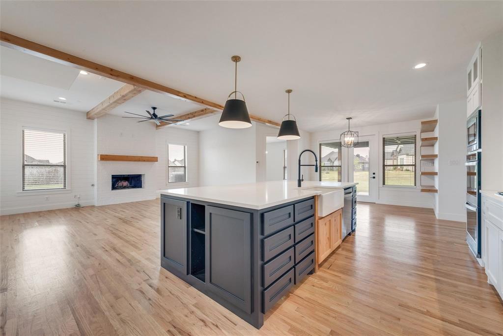 Open-concept kitchen featuring an island with stone-finish countertops, apron-front sink, and custom cabinetry, complemented by wood-finish flooring, exposed ceiling beams, and a fireplace with a wood mantel