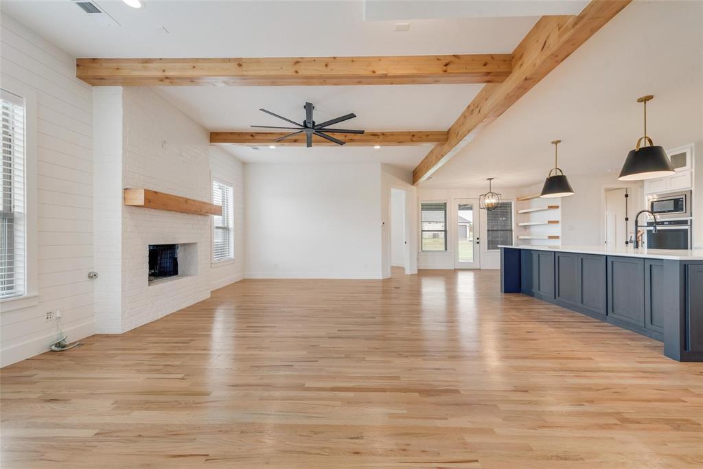 Open-concept interior featuring exposed wooden ceiling beams, wood-finish flooring, a white brick fireplace with a wood mantel, and kitchen island with built-in cabinetry and pendant lighting