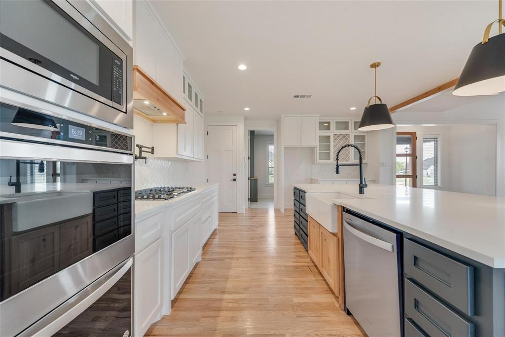 Kitchen featuring an expansive stone-surface island with a farmhouse sink, stainless steel wall-mounted oven and microwave, natural wood-finish flooring, white shaker-style cabinetry, and matte black pendant lighting