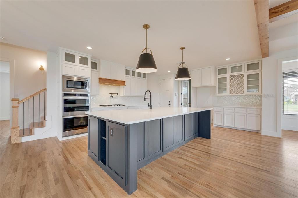 Kitchen featuring a central island with a solid surface countertop, two-toned cabinetry, built-in double wall ovens, glass-front upper cabinets with integrated wine storage, and wood-finish flooring Kitchen featuring a central island with a solid surface countertop, two-toned cabinetry, built-in double wall ovens, glass-front upper cabinets with integrated wine storage, and wood-finish flooring