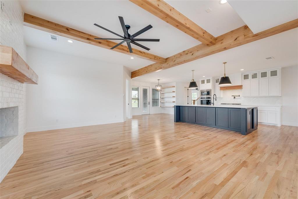 Open-concept interior featuring wood-finish flooring, exposed wooden ceiling beams, a painted brick fireplace with a wooden mantel, a large kitchen island with contrasting cabinetry, and floating wall shelves