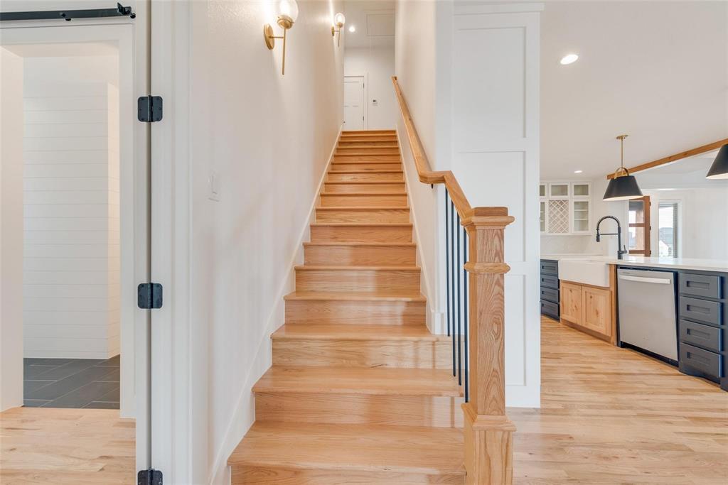 Staircase featuring wood-finish treads and a handrail with black metal balusters, complemented by wall-mounted sconce lighting, wood-finish flooring, and shaker-style cabinetry with a farmhouse-style sink