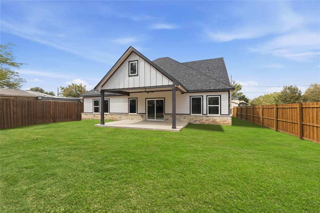 Rear exterior featuring a covered concrete patio, board-and-batten siding, stone masonry skirting, asphalt shingle roofing, and perimeter wood privacy fencing