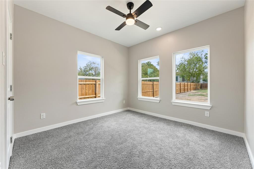 Neutral-toned interior featuring plush carpeting, three light-filled windows, and a matte black ceiling fan with integrated lighting