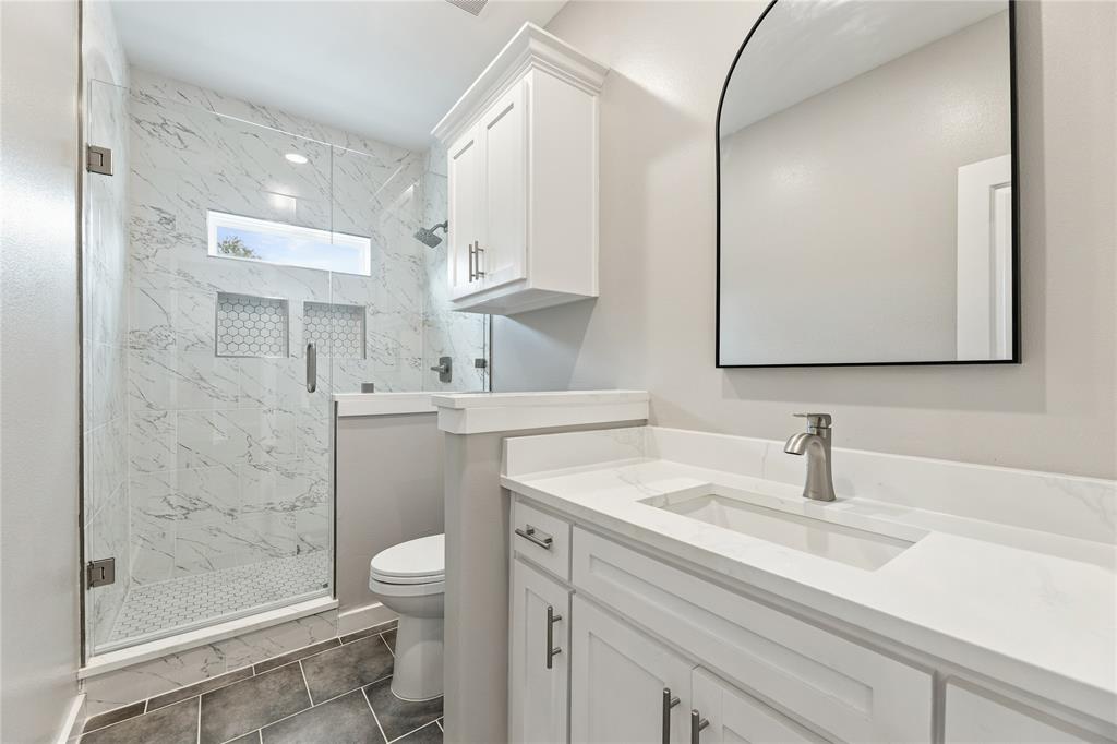 Walk-in shower with stone-finish tile surround and built-in niches, white cabinetry with countertop-integrated sink, arched wall mirror, and grey tile flooring