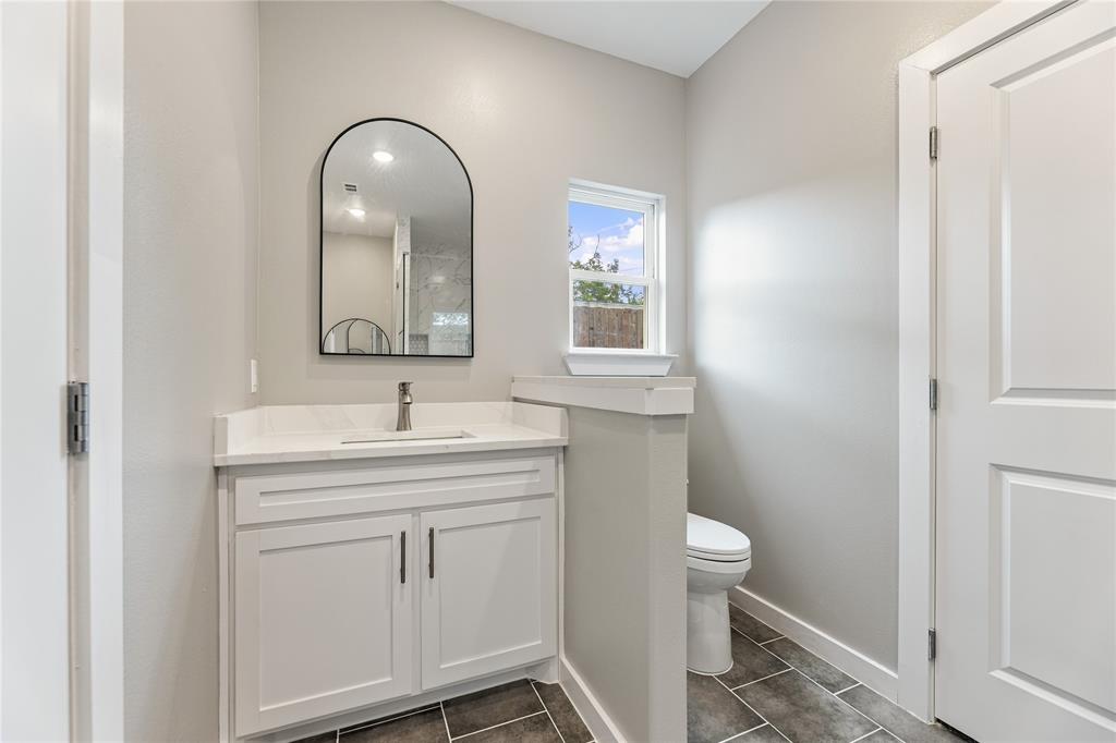 Bathroom featuring white shaker-style cabinetry, white stone-composite countertop with integrated sink, arched wall mirror, and porcelain tile flooring