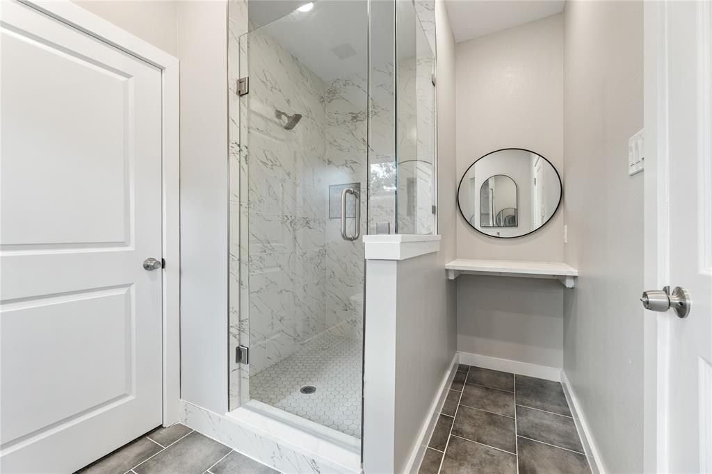 Walk-in shower with floor-to-ceiling stone-look tile, glass enclosure, hexagonal mosaic tile flooring, built-in shelving, and circular wall-mounted mirror