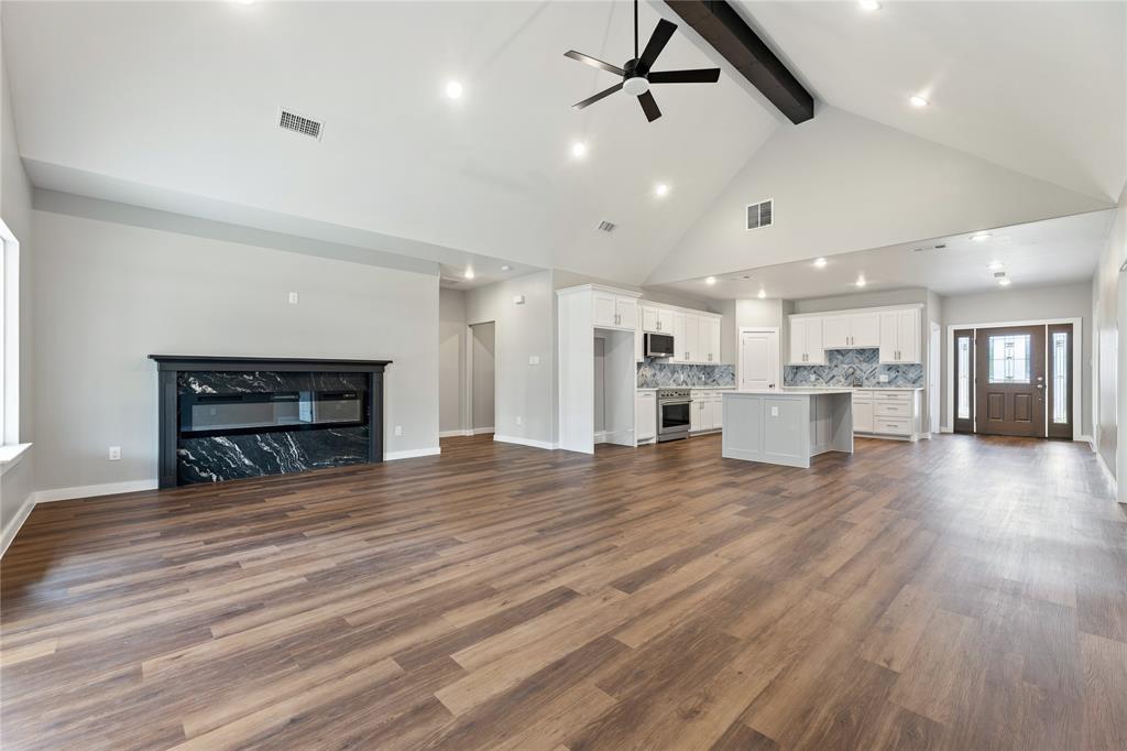 Vaulted ceilings with exposed structural beam, wood-finish flooring, stone-surround fireplace, kitchen with white cabinetry and central island, and recessed lighting