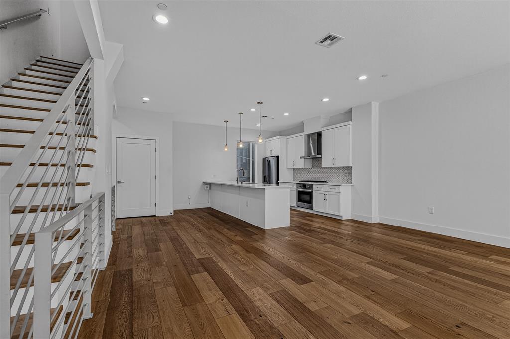 Open-concept interior featuring wood-finish flooring, recessed lighting, and a kitchen equipped with white cabinetry, a center island, and stainless steel appliances, accented by a staircase with horizontal metal railing