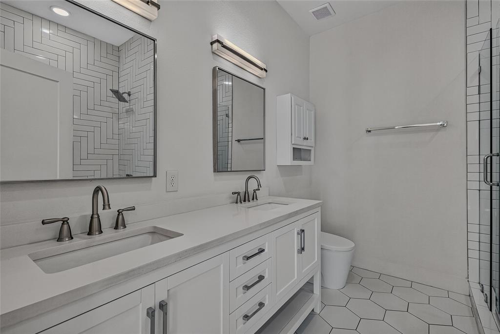 Dual-sink vanity with quartz countertop, white shaker-style cabinetry, matte black hardware, glass-enclosed shower with herringbone tile detailing, and hexagonal floor tiling