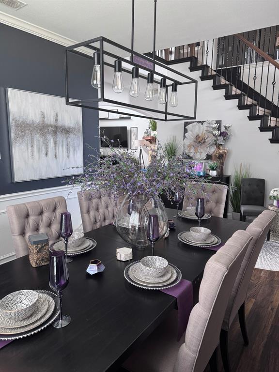 Dining area featuring a linear matte black chandelier, dark accent wall with chair rail molding, and an open staircase with black metal balusters and wood-finish treads Dining area featuring a linear matte black chandelier, dark accent wall with chair rail molding, and an open staircase with black metal balusters and wood-finish treads