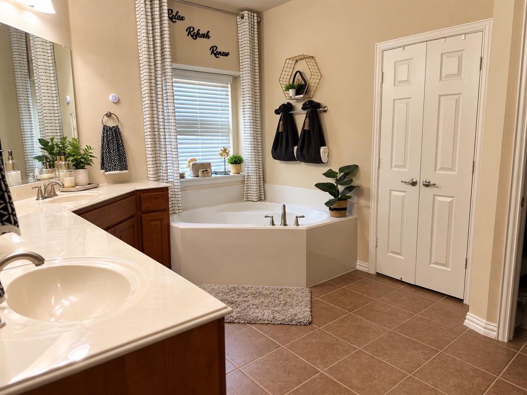 Bathroom featuring a double-sink vanity with light-toned countertops, a corner soaking tub with chrome fixtures, and tile flooring Bathroom featuring a double-sink vanity with light-toned countertops, a corner soaking tub with chrome fixtures, and tile flooring