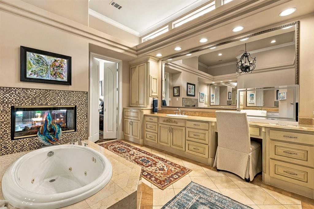 Jetted soaking tub with tile surround, double-sided fireplace with mosaic tile detailing, dual-sink vanity with custom cabinetry, expansive wall-mounted mirror, and recessed lighting