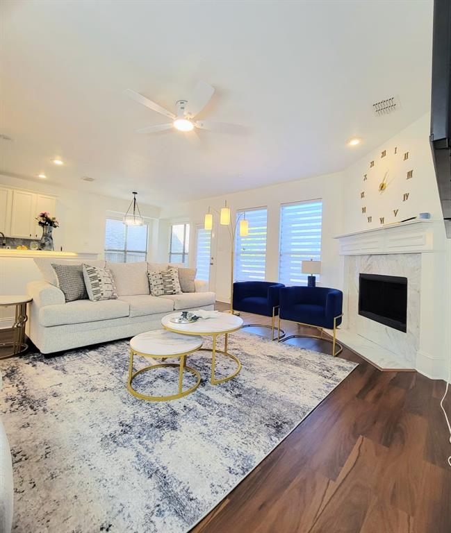 Living area featuring a fireplace with a white mantel and stone-finish surround, wood-finish flooring, recessed lighting, and a ceiling fan Living area featuring a fireplace with a white mantel and stone-finish surround, wood-finish flooring, recessed lighting, and a ceiling fan