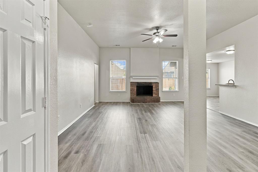 Open-concept room featuring wood-finish flooring, a brick fireplace with a mantel, dual windows, and a ceiling fan with integrated lighting Open-concept room featuring wood-finish flooring, a brick fireplace with a mantel, dual windows, and a ceiling fan with integrated lighting