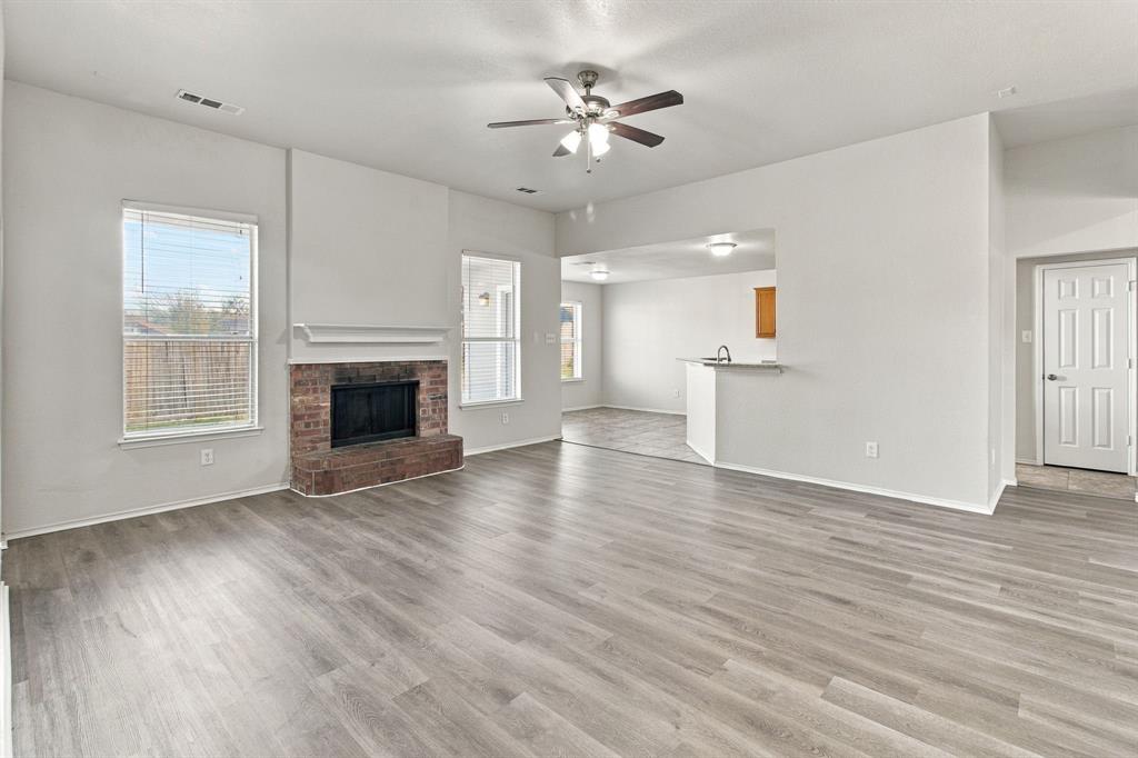 Open-concept living space featuring wood-finish flooring, a brick fireplace with a mantle, dual-aspect windows, and a ceiling fan with integrated lighting