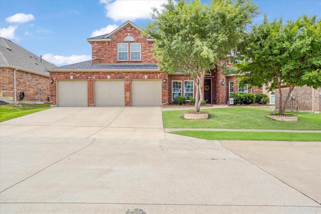 Two-story brick exterior featuring a three-car garage, concrete driveway, and established front landscaping with mature trees and stone-ringed planting beds