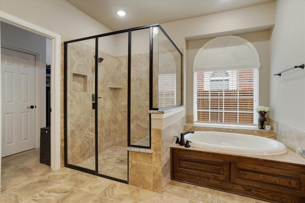 Bathroom featuring a frameless glass shower enclosure with dark bronze accents, a built-in soaking tub with dark wood paneling, and a large arched window with blinds