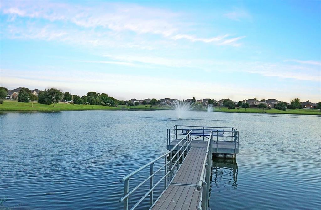 Floating dock with metal railings extending into a body of water featuring two active fountains, surrounded by a manicured green shoreline and residential properties