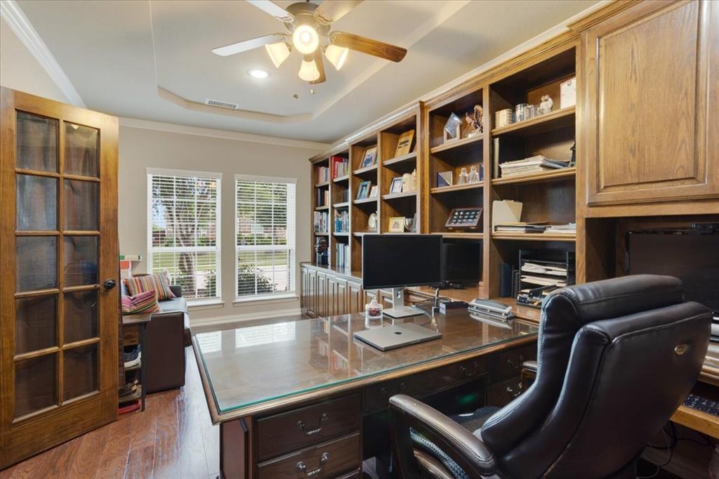 Dedicated office space featuring extensive built-in wood cabinetry and shelving, a tray ceiling with a ceiling fan, wood-finish flooring, and a glass-paneled entry door