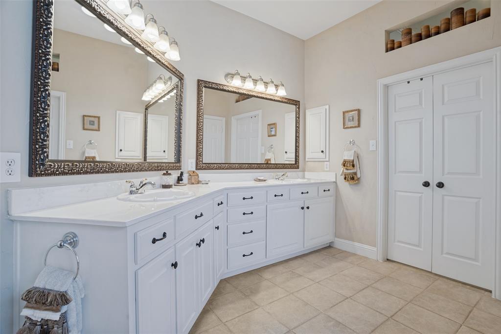 Dual-sink vanity with white cabinetry, light-toned countertop, and decorative framed mirrors, complemented by ceramic tile flooring and recessed wall shelving