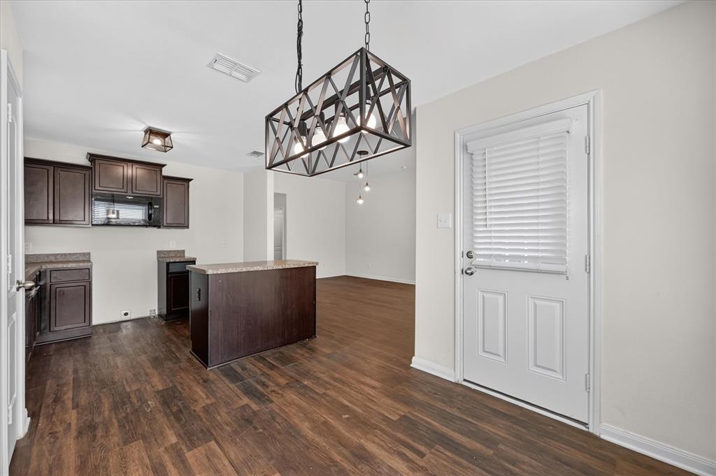 Dark-finish cabinetry, wood-finish flooring, central kitchen island, geometric metal-frame pendant lighting, and an exterior door with integrated blinds Dark-finish cabinetry, wood-finish flooring, central kitchen island, geometric metal-frame pendant lighting, and an exterior door with integrated blinds