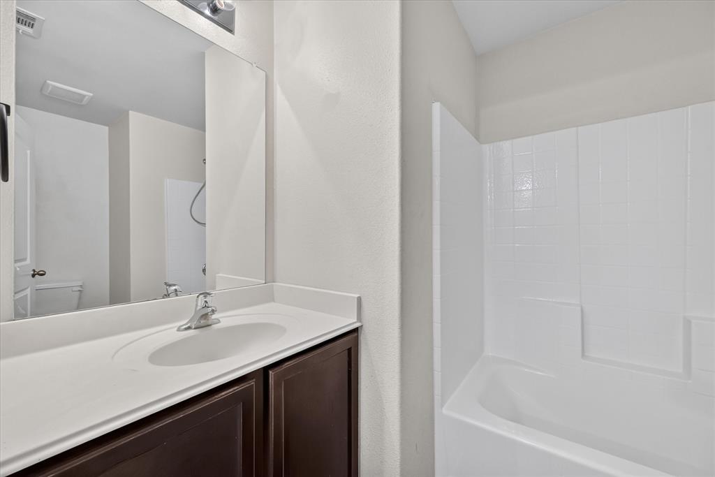 Bathroom featuring a dark wood-finish vanity, integrated white countertop with a single-basin sink, polished chrome faucet, full-width mirror, and a white tiled bathtub surround