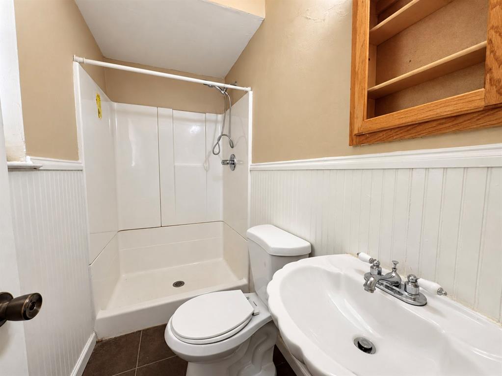 Bathroom featuring a white molded shower surround, white beadboard wainscoting, a wall-mounted wood-finish medicine cabinet, and a scalloped porcelain sink with a polished chrome faucet