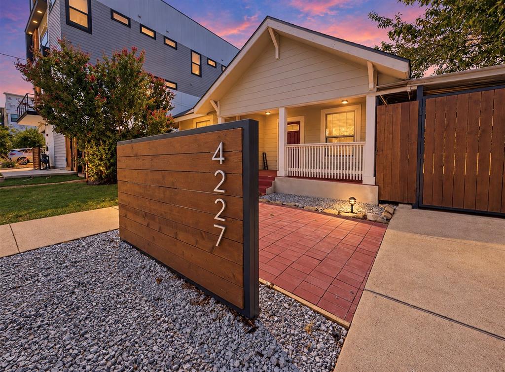 Exterior facade featuring horizontal wood-plank accent wall, paved brick patio, covered front porch with railing, and gravel landscaping