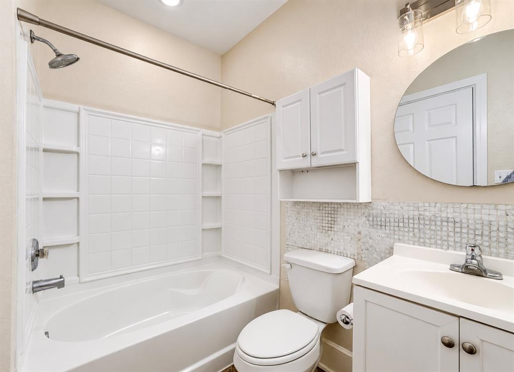 Bathroom featuring a white tub-and-shower combination with integrated shelving, white vanity cabinet with a solid-surface top, circular wall-mounted mirror, and wall-mounted storage cabinetry