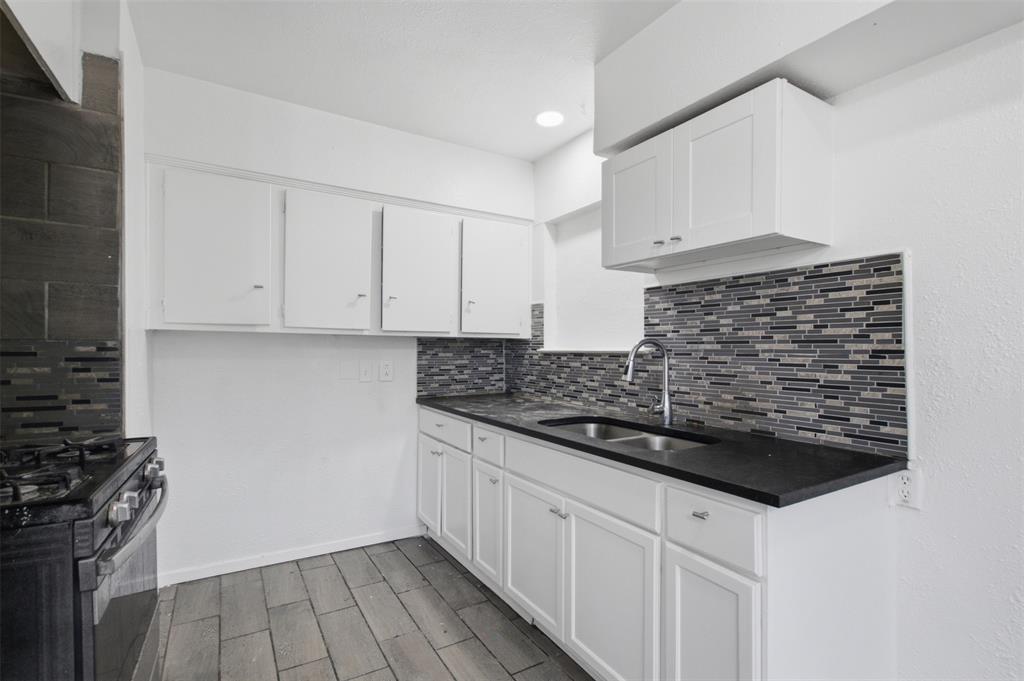 Kitchen featuring white cabinetry, dark countertops, a multi-tone linear tile backsplash, stainless steel double-basin sink with pull-down faucet, and wood-finish flooring Kitchen featuring white cabinetry, dark countertops, a multi-tone linear tile backsplash, stainless steel double-basin sink with pull-down faucet, and wood-finish flooring