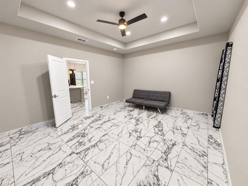 Tray ceiling with integrated recessed lighting, large-format veined tile flooring, and a central matte black ceiling fan