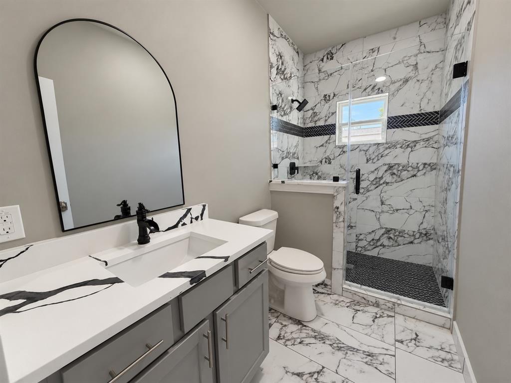 Bathroom featuring a gray vanity with a stone-finish countertop, arched-top mirror, walk-in shower with large-format tile surrounds, and matte black fixtures