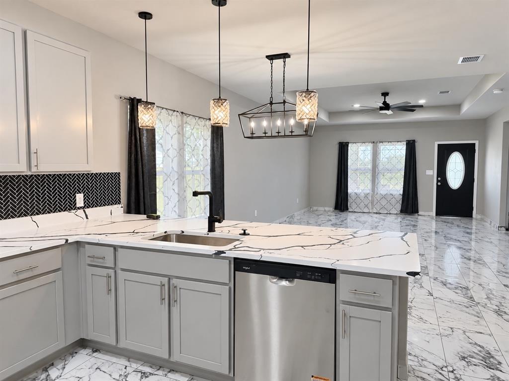 Kitchen featuring light gray shaker-style cabinetry, quartz countertops with prominent veining, a black herringbone tile backsplash, stainless steel dishwasher, and polished stone-finish flooring Kitchen featuring light gray shaker-style cabinetry, quartz countertops with prominent veining, a black herringbone tile backsplash, stainless steel dishwasher, and polished stone-finish flooring